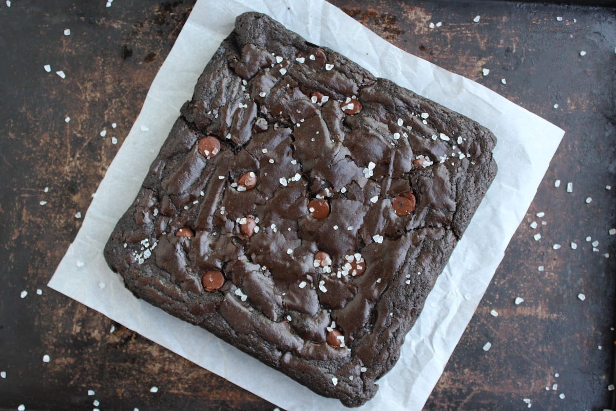 uncut batch of brownies on parchment paper, topped with flaky salt and chocolate chips