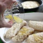 A hand holding a small glass of melted better, which is being poured over breaded fillets of haddock in a white baking dish