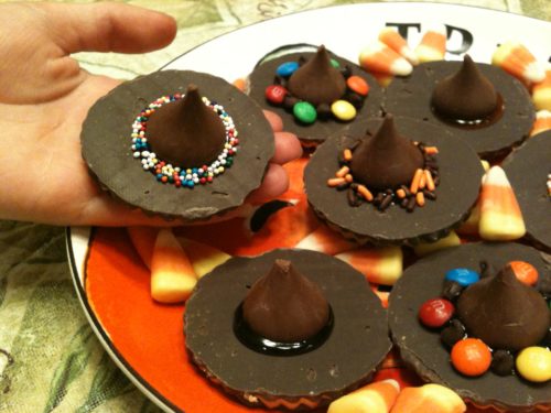 A child's hand holding a choclate witch hat cookie, above a plateful of cookies.