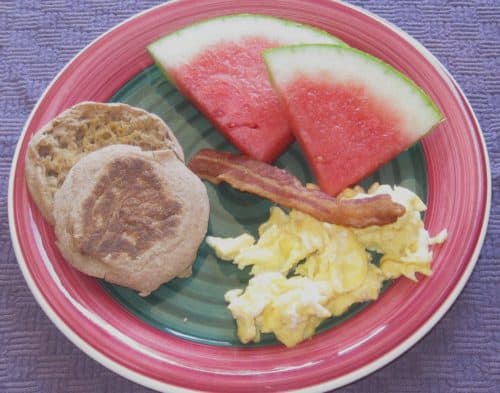 Plate of scrambled eggs with bacon, watermelon, and whole grain english muffin